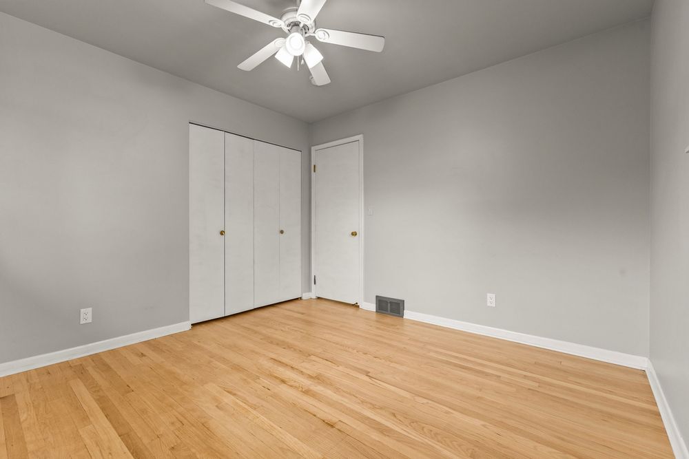 Bright empty bedroom with hardwood floors, ceiling fan, and white closet doors.