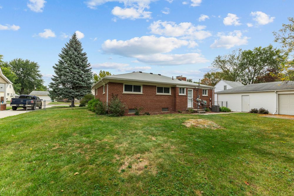 Rear view of brick ranch home with spacious green backyard and detached garage on a sunny day.