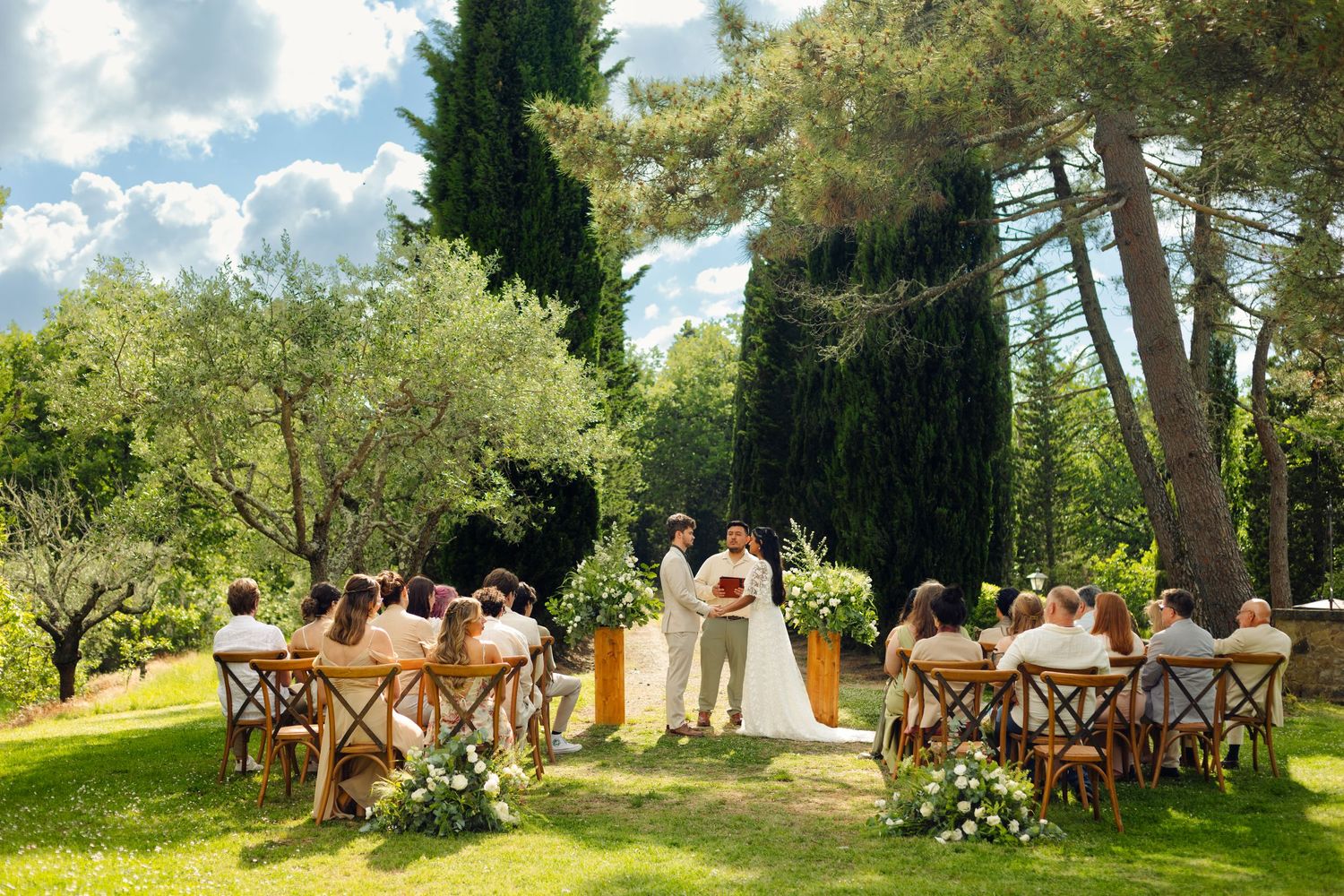 Garden wedding ceremony setup with wooden chairs and floral arrangements surrounded by cypress trees.