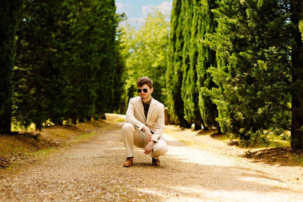 Person in beige suit crouches on gravel path surrounded by towering cypress trees.