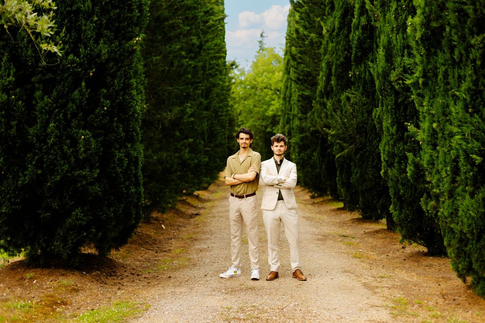 Two people in light colored suits stand on path lined with tall cypress trees.