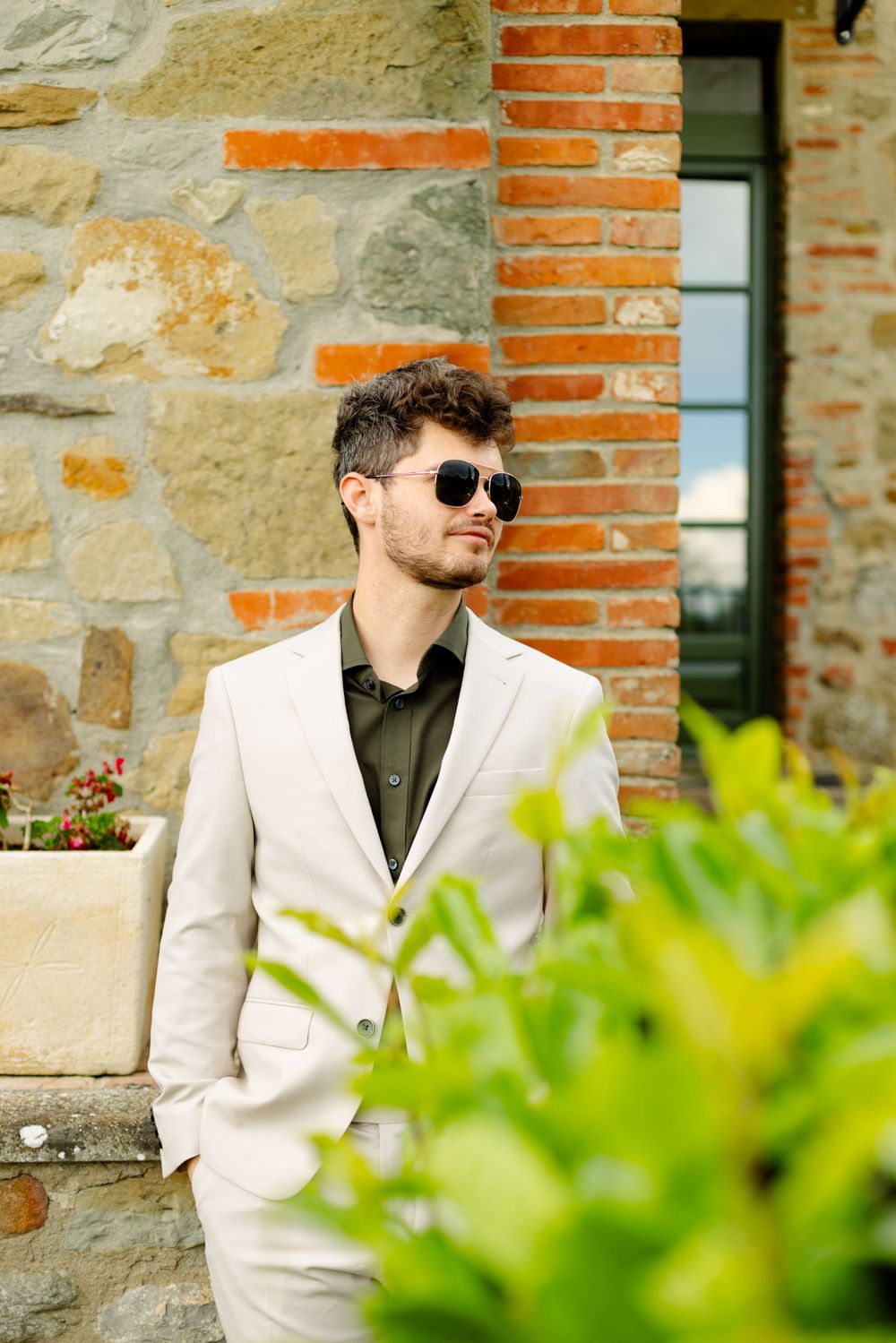 Person in cream suit and sunglasses poses against stone and brick wall with green foliage.
