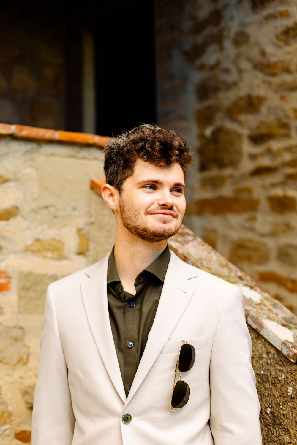 Person in cream suit and dark shirt smiles while standing against stone wall backdrop.
