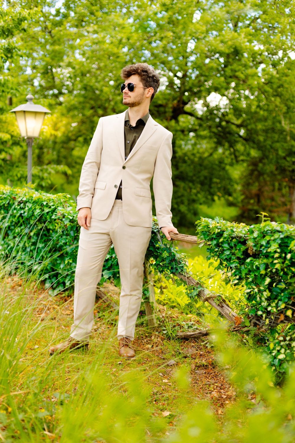Person in beige suit and sunglasses strolls through lush garden setting with lamp post.