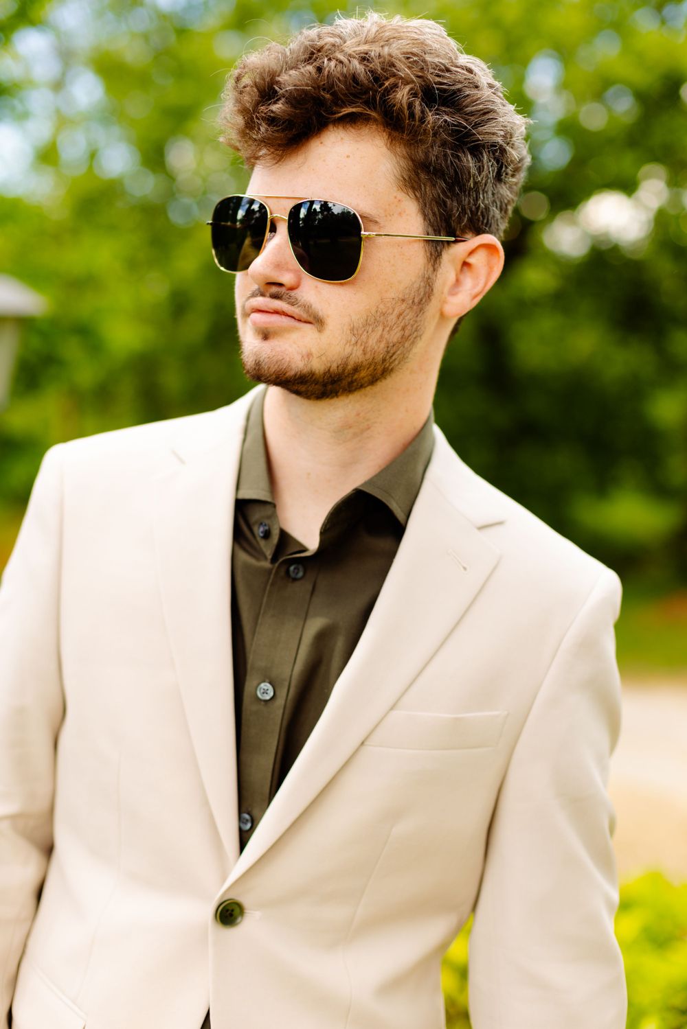 Person in cream blazer and dark shirt wearing aviator sunglasses against blurred green outdoor background.