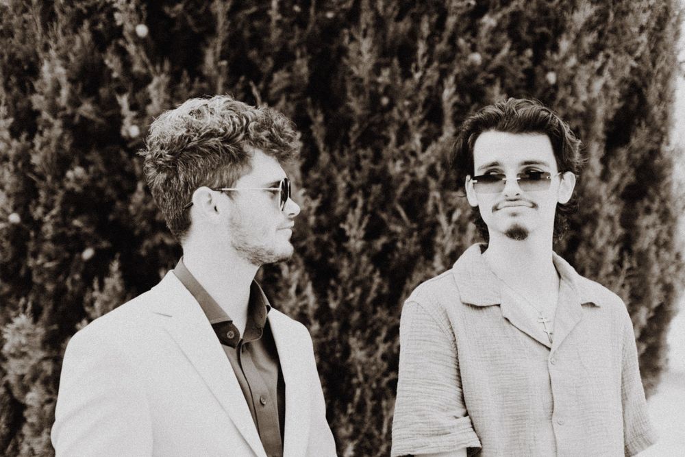 Black and white portrait of two people in formal attire standing against foliage backdrop.