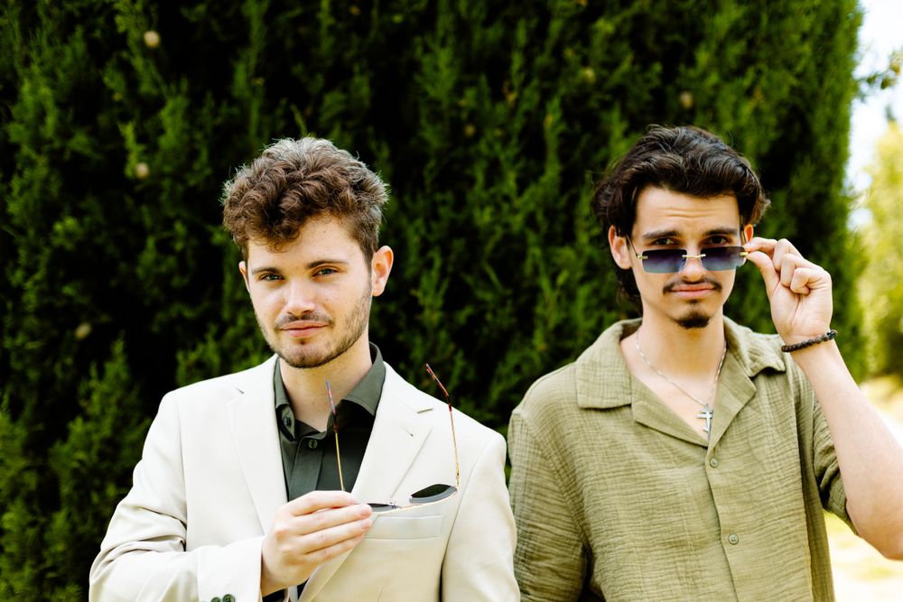 Two stylishly dressed people stand outdoors with sunglasses against greenery in the background.
