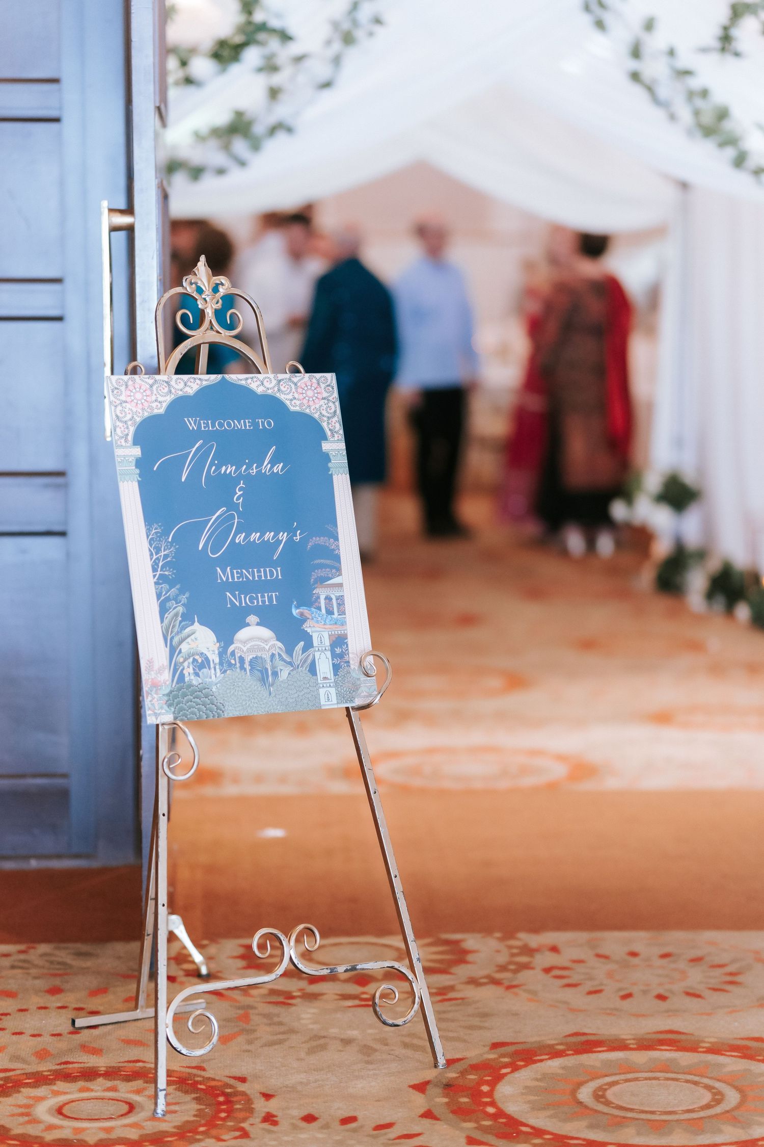 Ornate blue and white wedding seating cards displayed on decorative metal stands against a red patterned carpet.