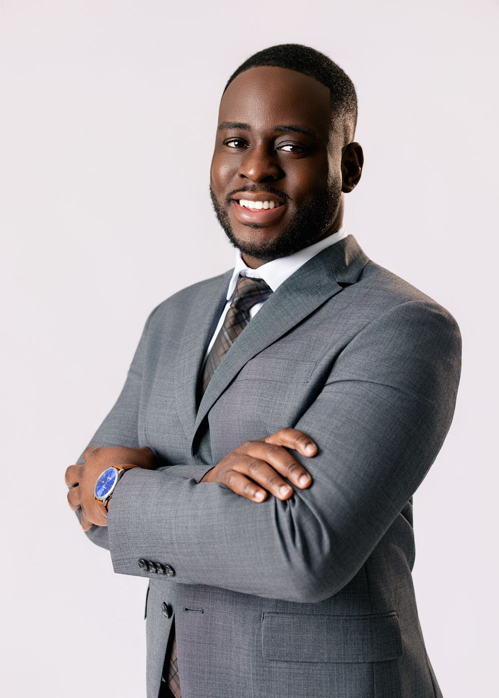 Professional headshot of someone wearing a grey suit and patterned tie posing with arms crossed against white background.