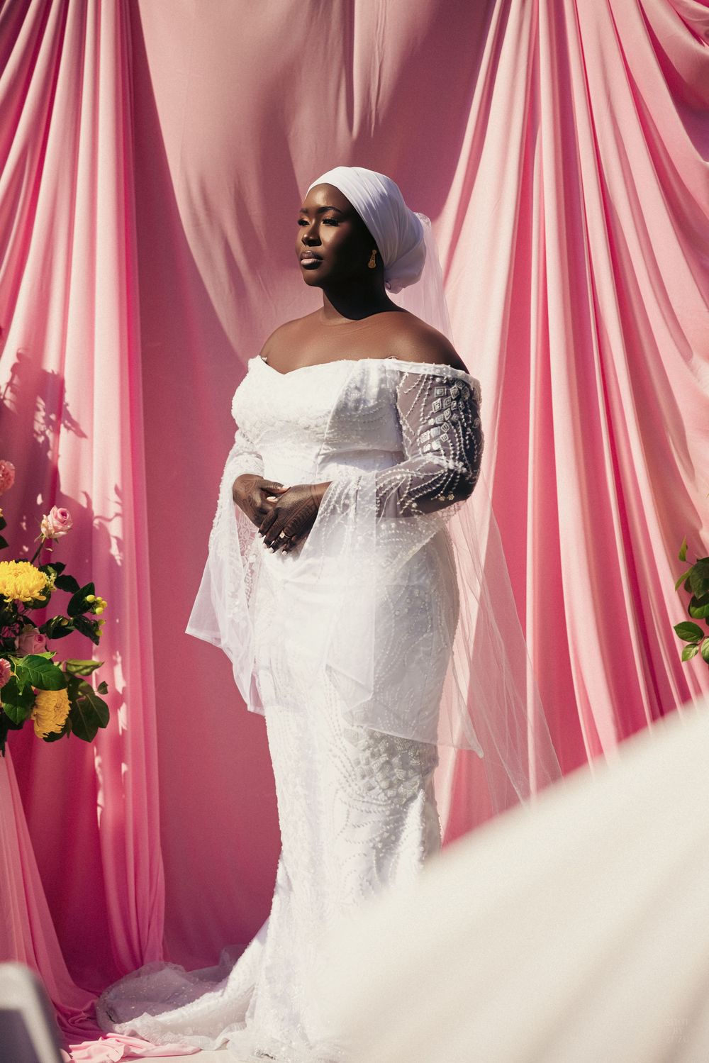 A bride wearing an elegant off-shoulder wedding gown poses against a pink draped backdrop.