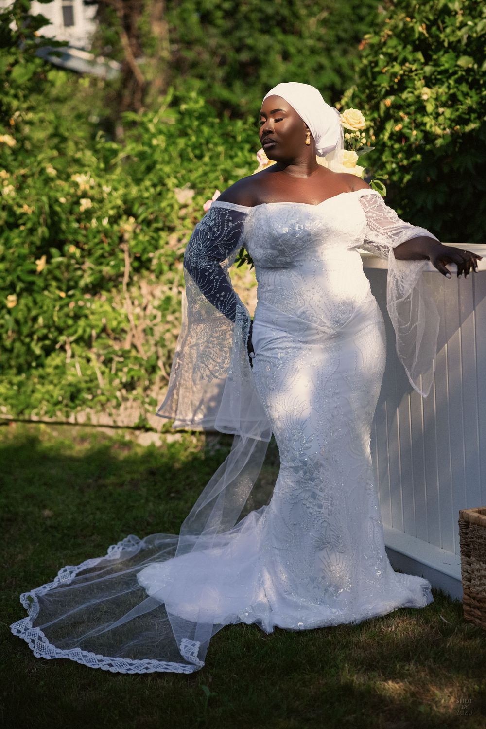 A bride in a white lace wedding dress poses gracefully outdoors against a garden backdrop with a flowing veil.