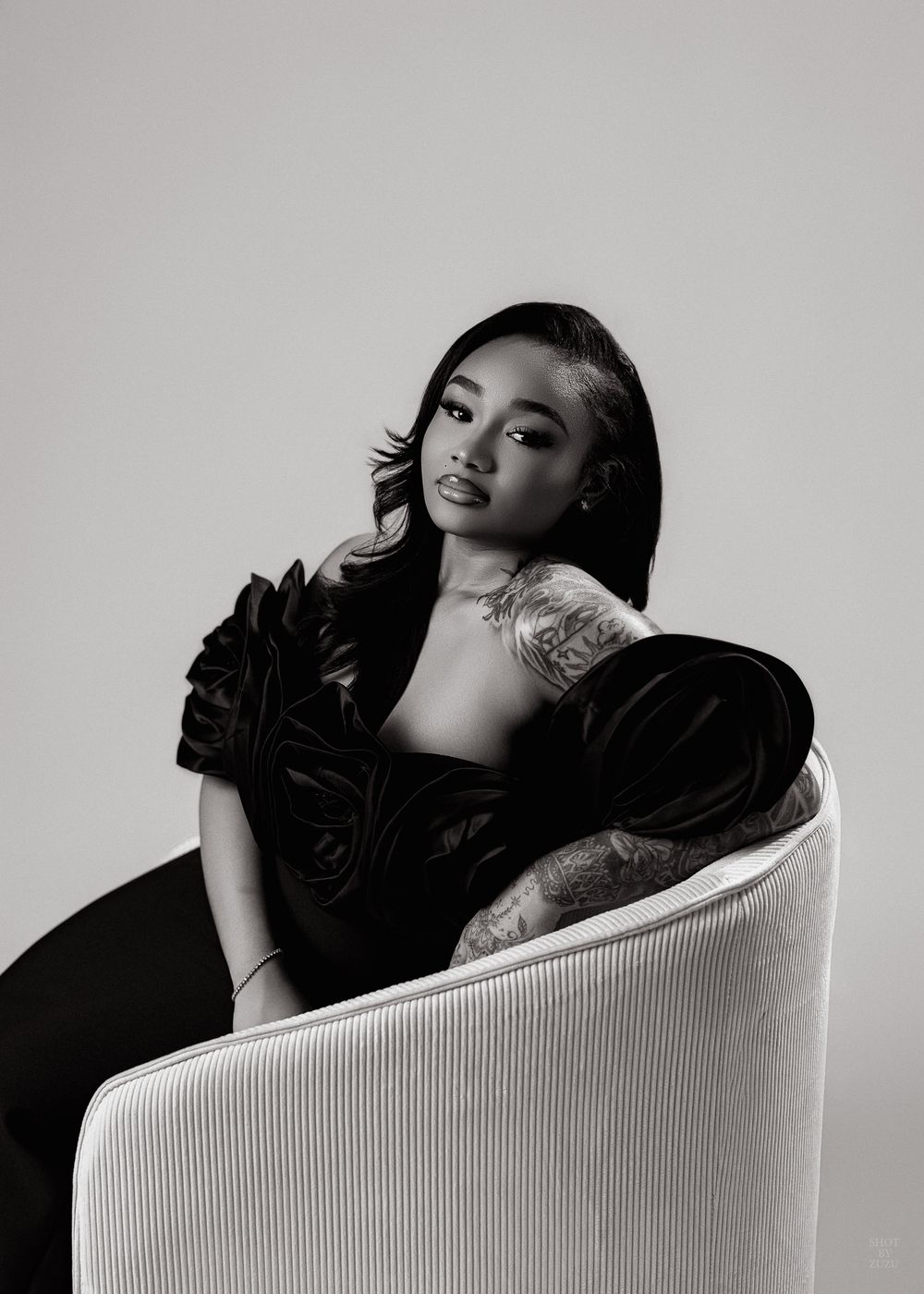 Artistic black and white portrait of someone in a dark velvet dress posing elegantly on a white ribbed chair.