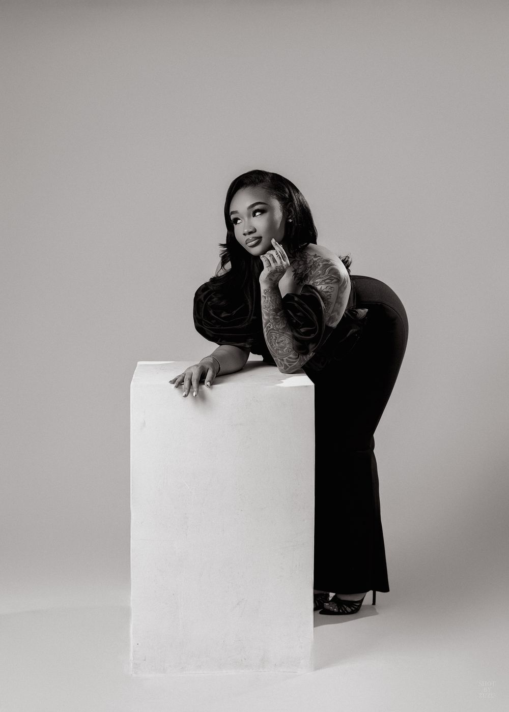 A black and white studio portrait of a person in a dark outfit posing elegantly against a white pedestal.