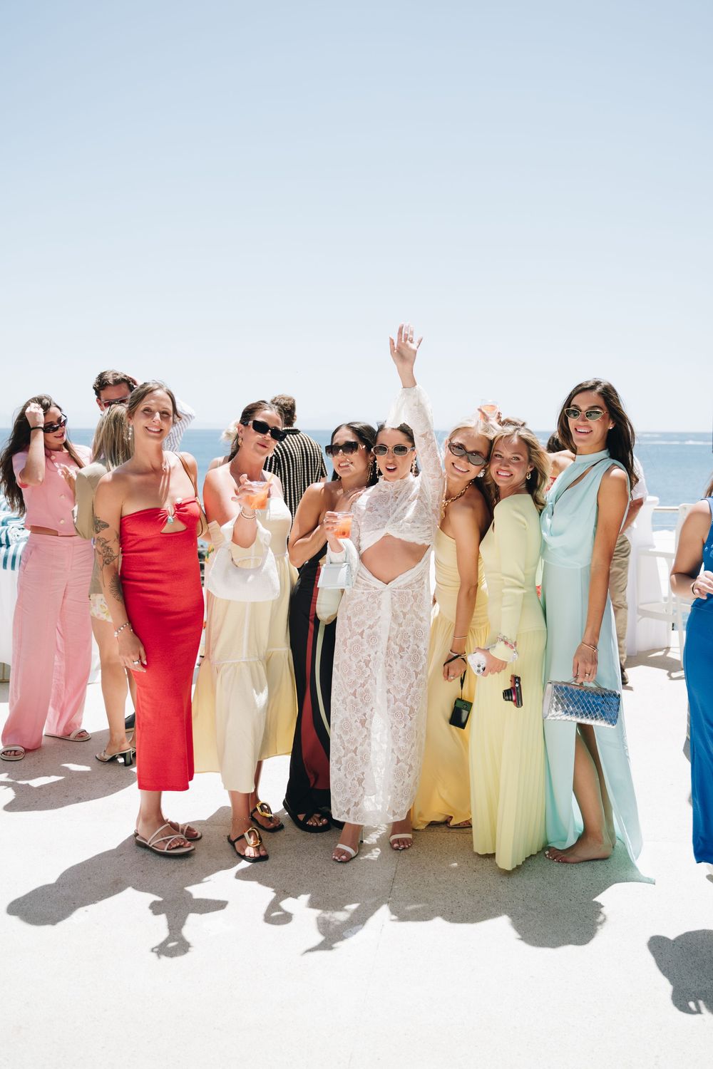 A group of friends in colorful summer dresses and jumpsuits pose together at a sunny outdoor celebration by the ocean.