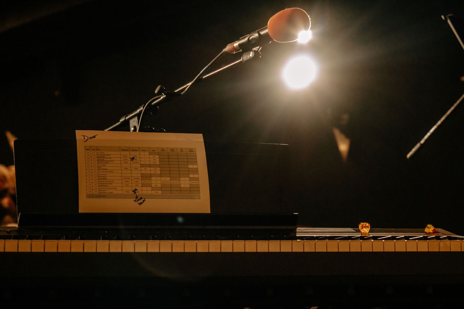 Piano keyboard illuminated by warm stage lighting with sheet music stand and microphone in dark performance setting.