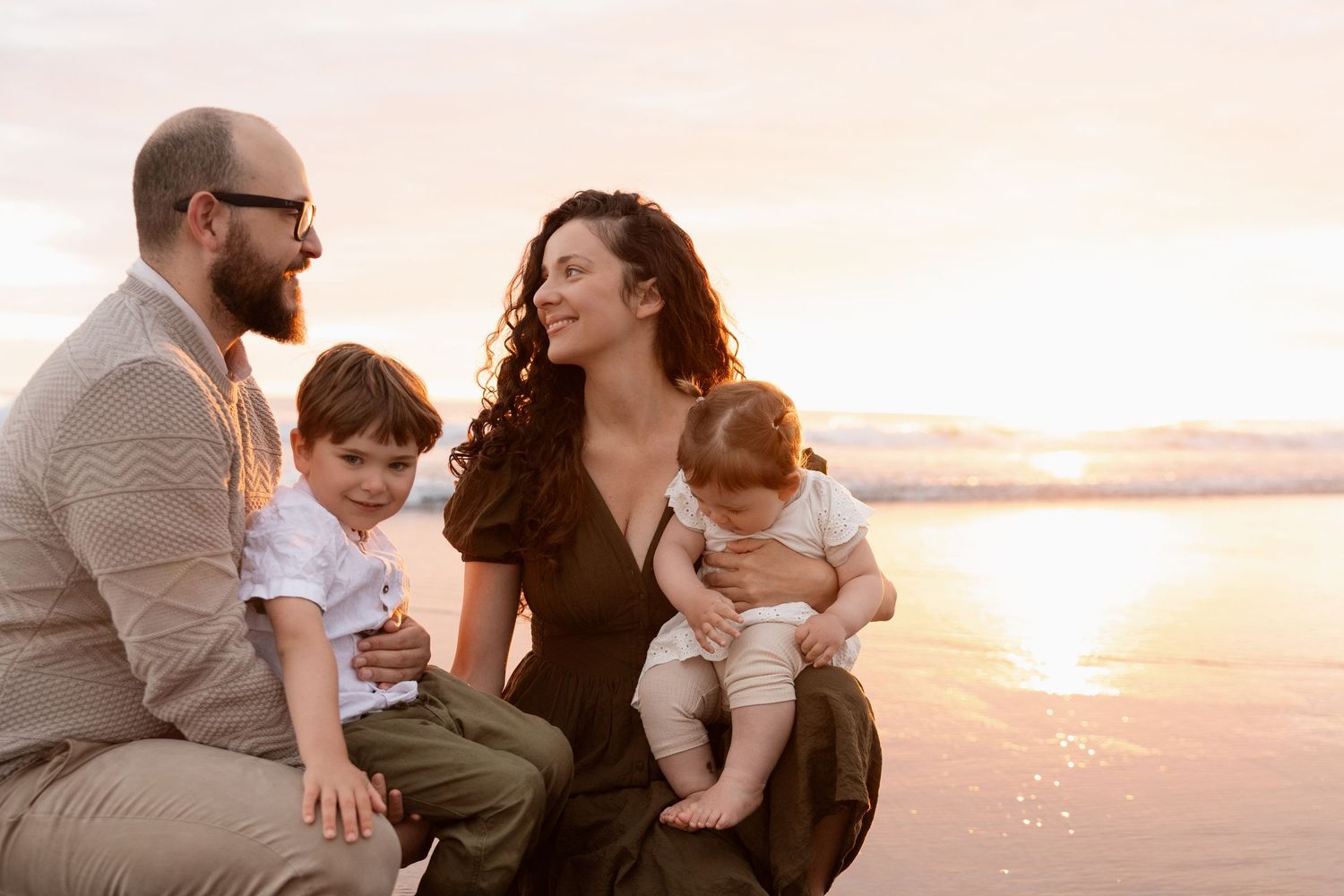 Mum holding daughter and dad holding son while looking at each other during sunrise beach session in Christchurch