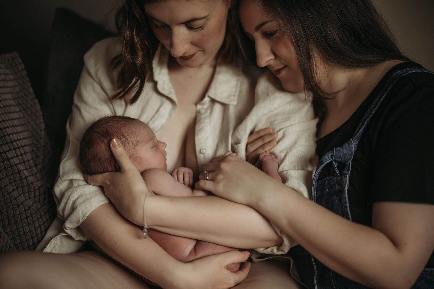 Two mothers sitting close together, gently holding their newborn baby in a quiet, intimate moment at home