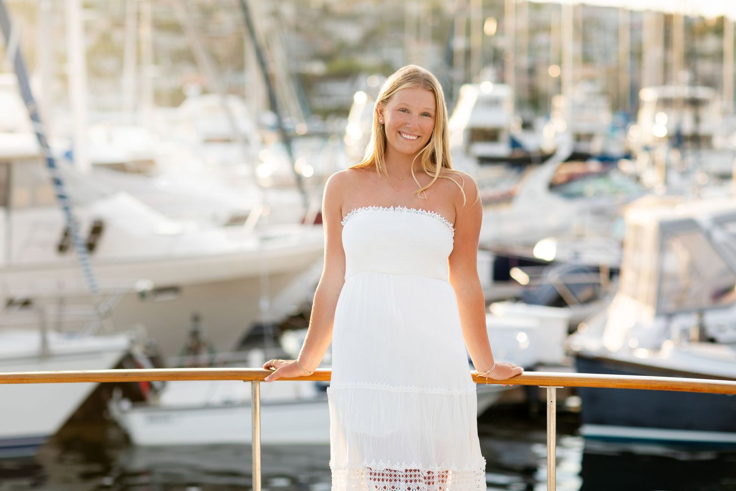 A high school senior in a white strapless dress stands at a marina with boats in the background on a sunny day.