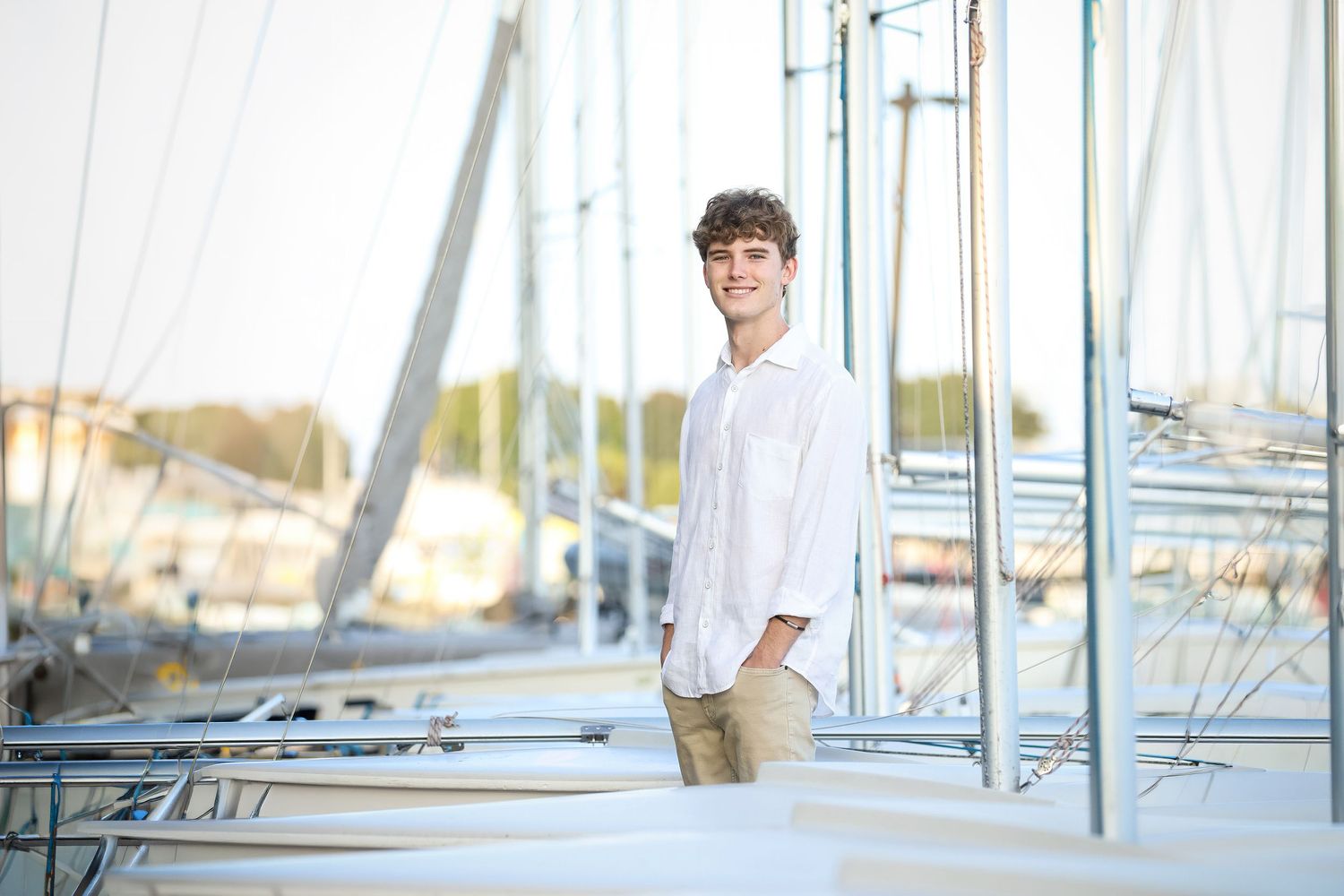 A high school senior in a white strapless dress stands at a marina with boats in the background on a sunny day.