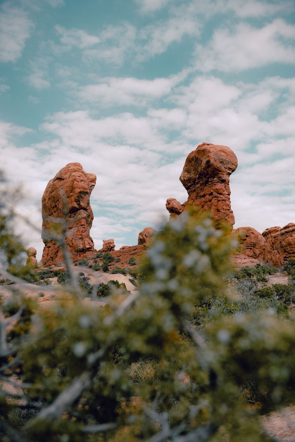 Natural rock formations rise against a cloudy blue sky with desert vegetation in the foreground at Arches National Park.