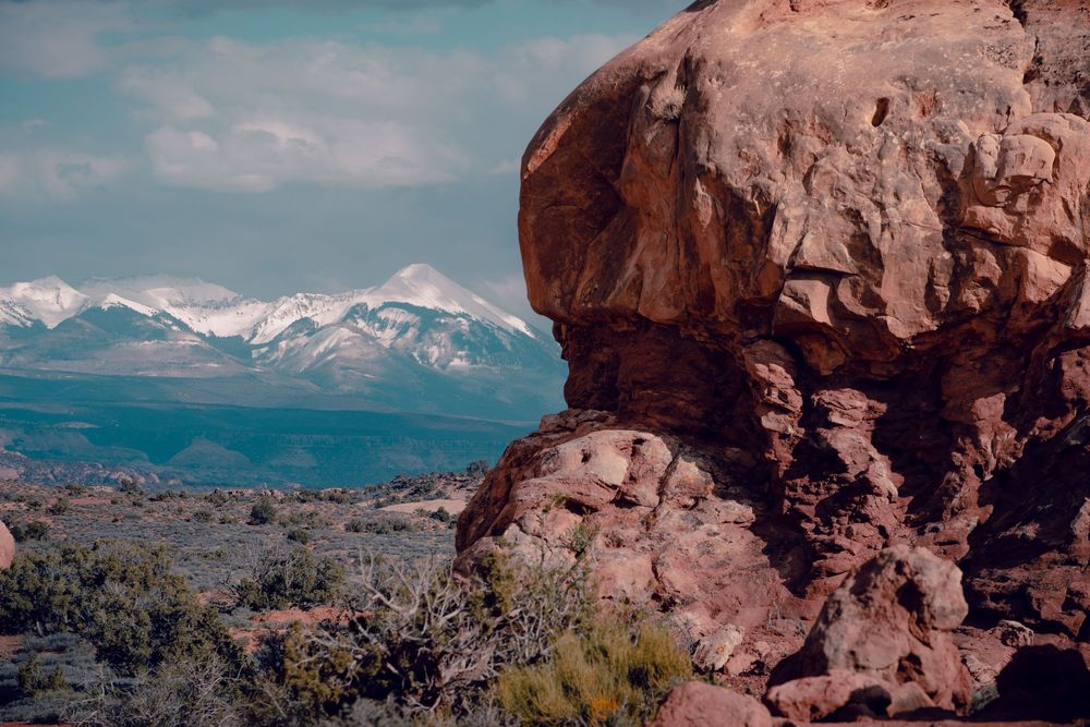 Large red rock formation in desert landscape with snow-capped mountains visible in the distance against blue sky.
