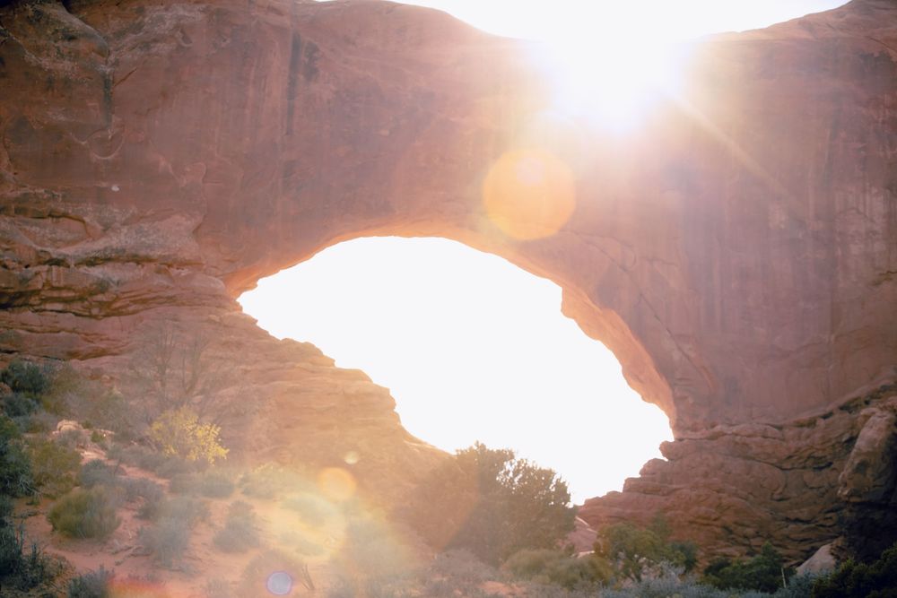 Sun rays shine through a natural rock arch formation in a desert canyon creating a dramatic lens flare effect.