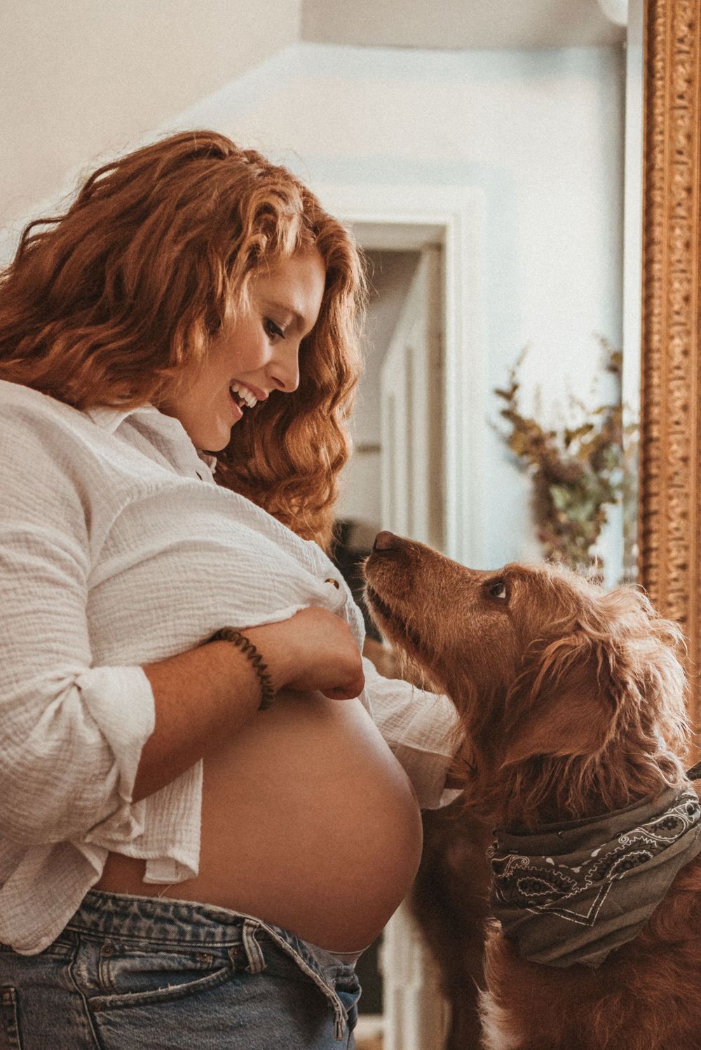 Pregnant woman in white shirt with dog showing affection to baby bump.