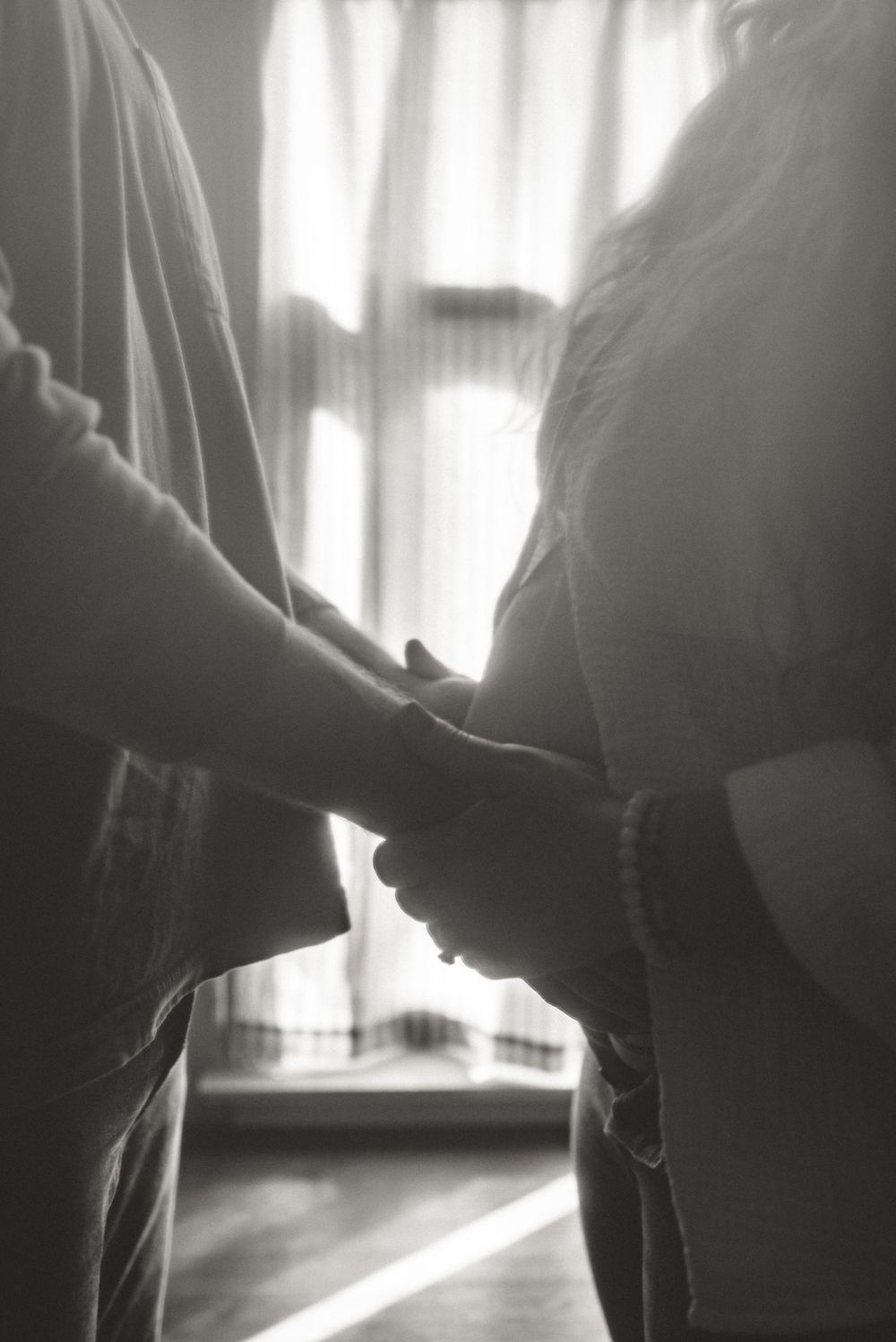 Black and white intimate silhouette of hands touching pregnant belly near window.