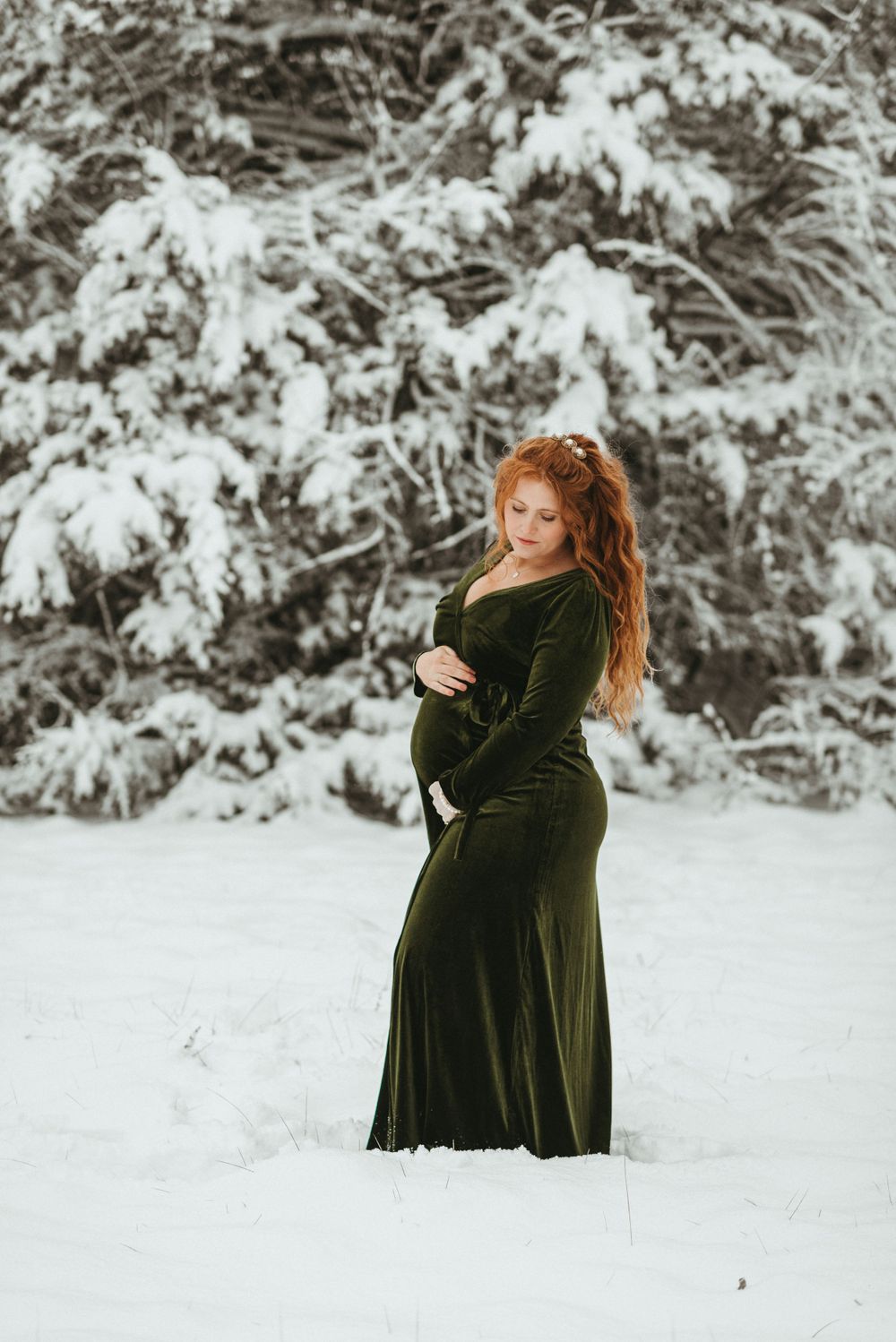Winter maternity photos with emerald velvet dress against snowy forest backdrop.