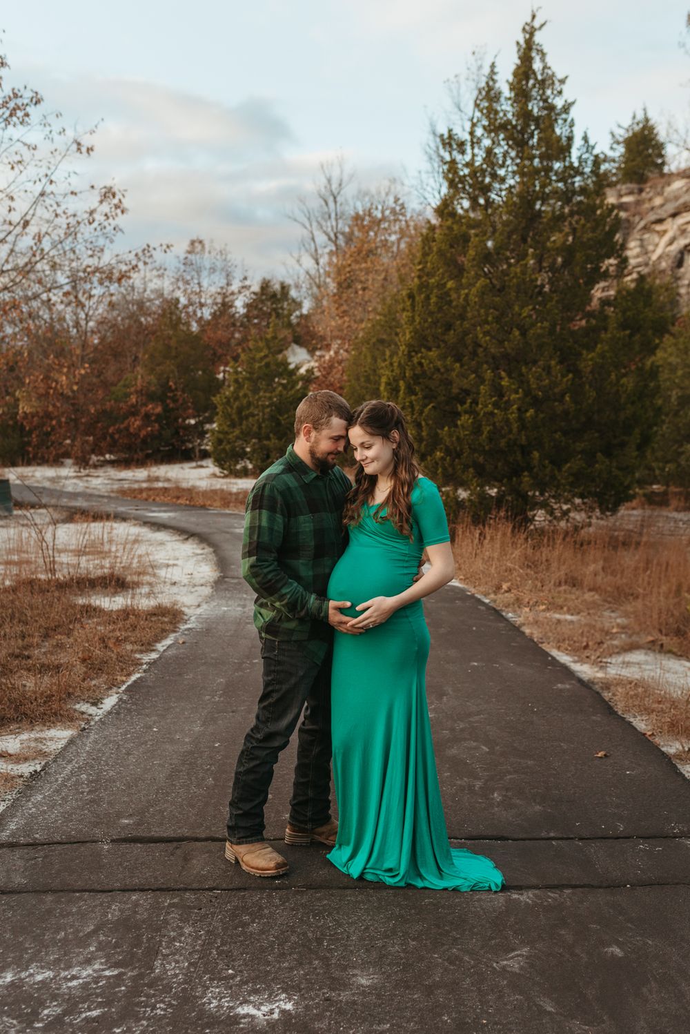 A romantic maternity photo on a path with fall foliage and evergreen trees.