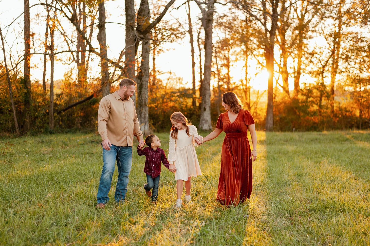 A joyful family walks through a golden sunlit field at sunset with two young children holding hands.