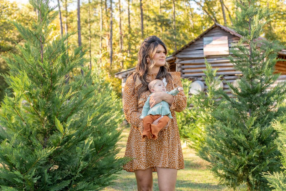 Mother and baby stand in front of old barn by Christmas Trees at Lakewood Tree Farm