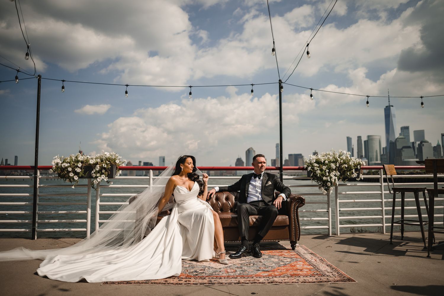 A wedding couple sits on a rooftop lounge area with string lights and the city skyline in the background.