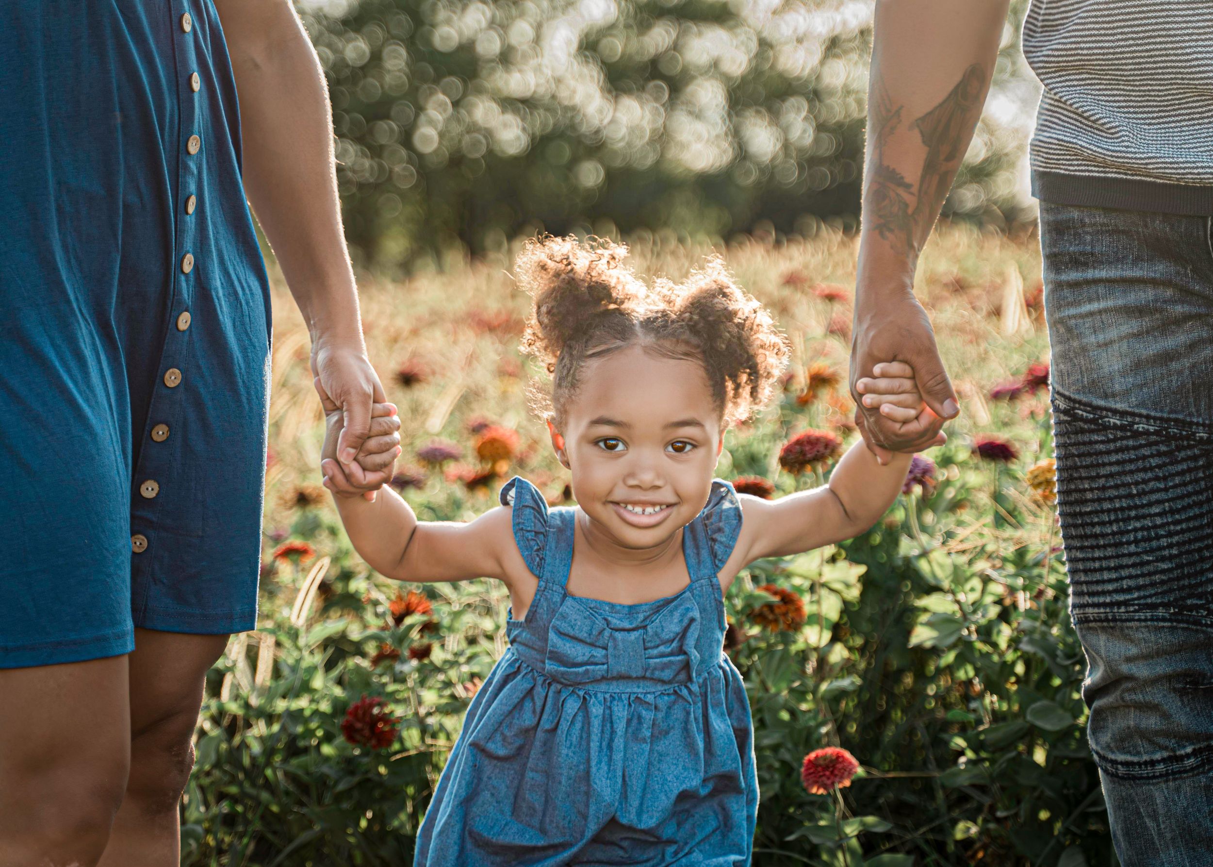 A joyful young child in a blue dress holds hands with adults while standing in a sunlit garden with flowers.