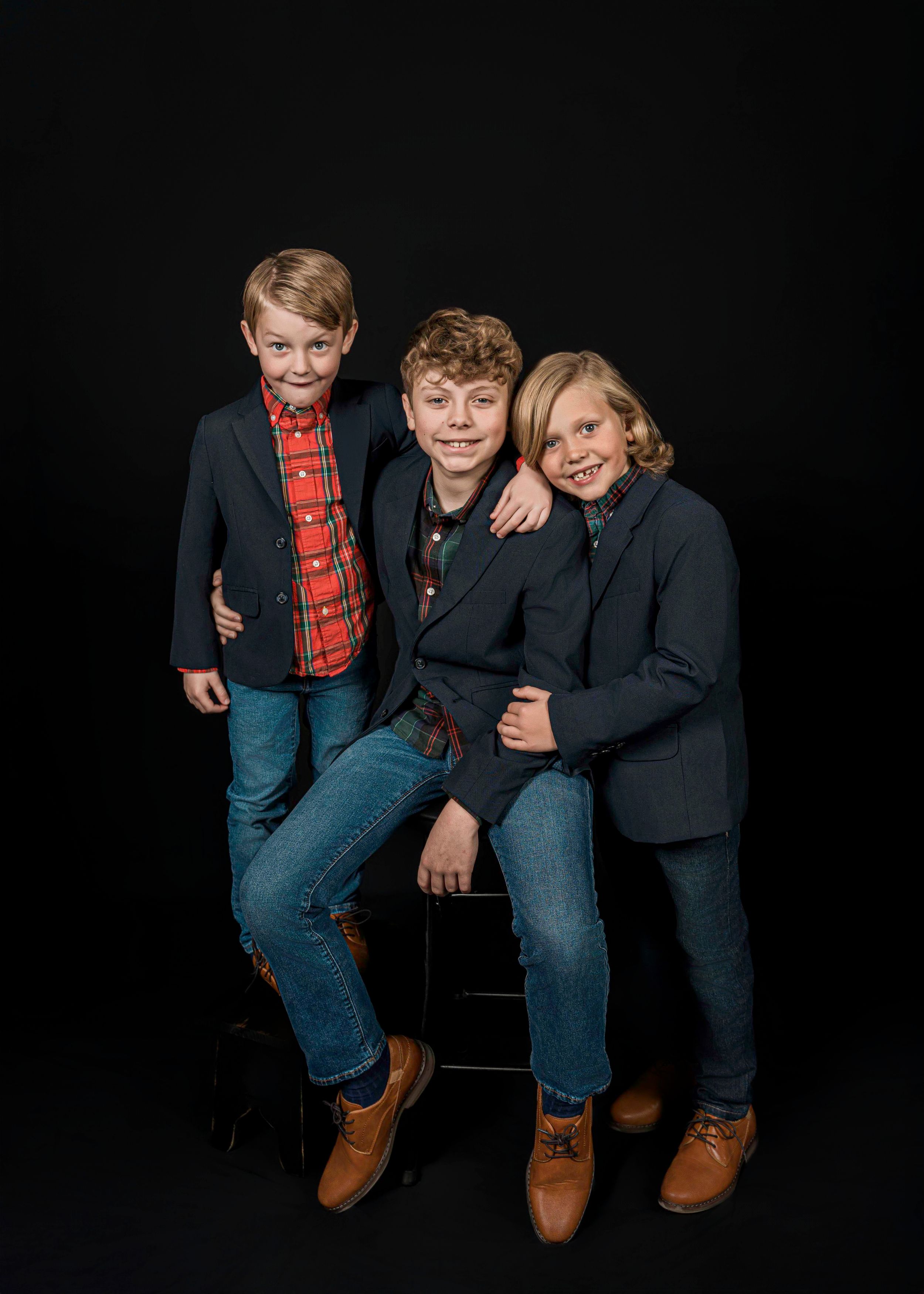 Three young people in blue jeans and blazers pose together against a black background in a stylish family portrait.