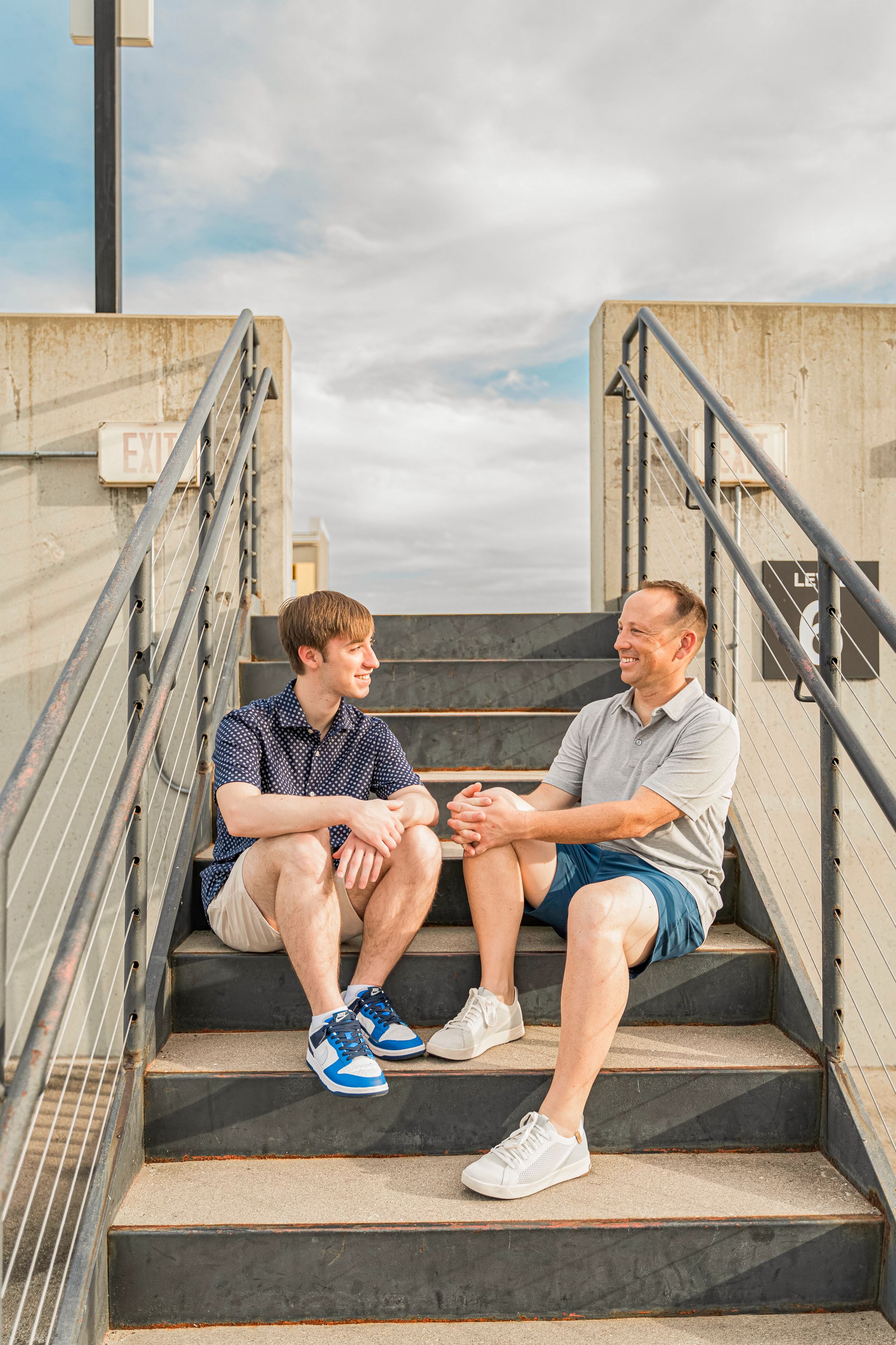 Two people sit having a conversation on concrete steps against a cloudy sky background.