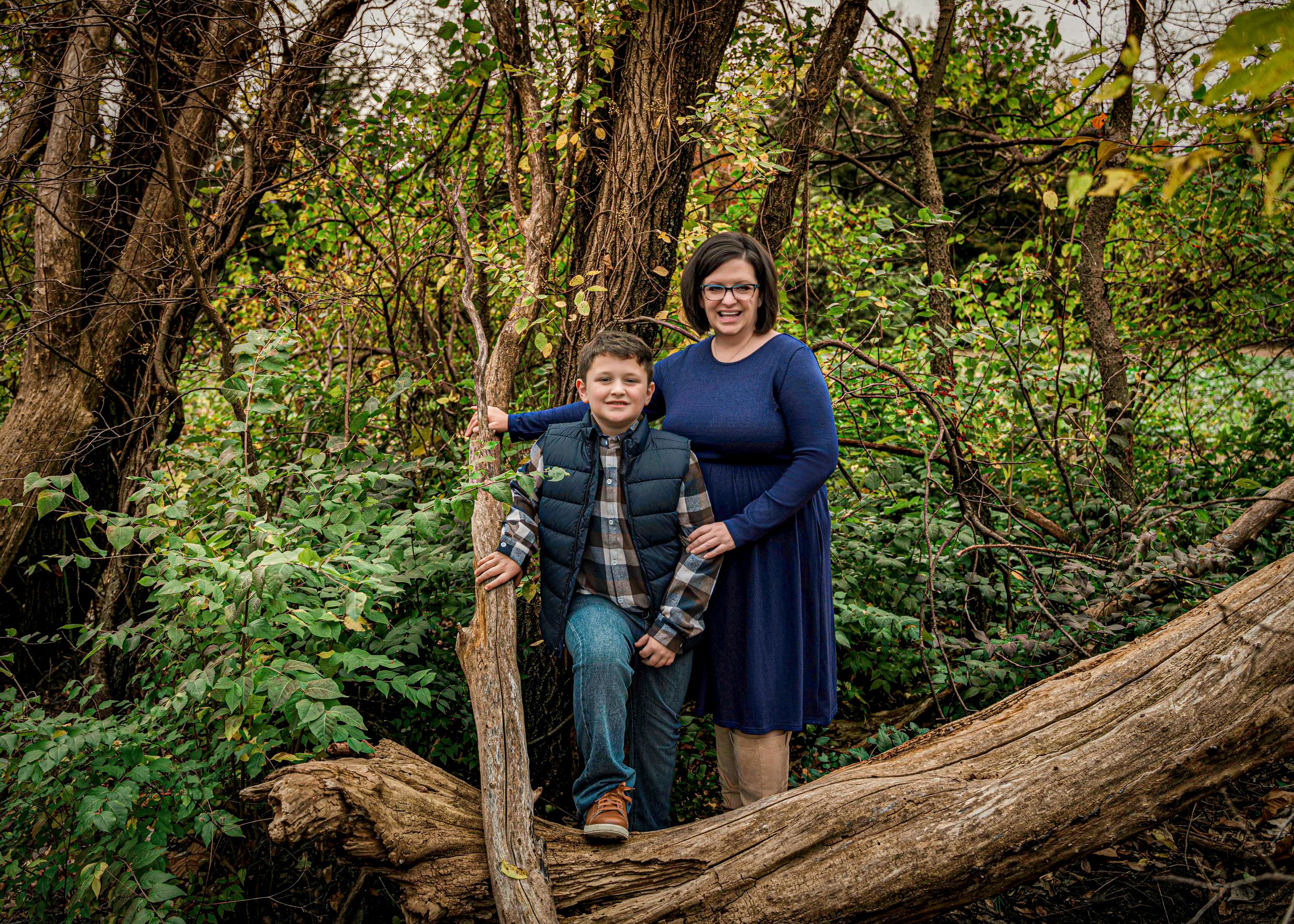 Mo.m and son wearing casual clothing pose together on a fallen log in a lush forest setting.