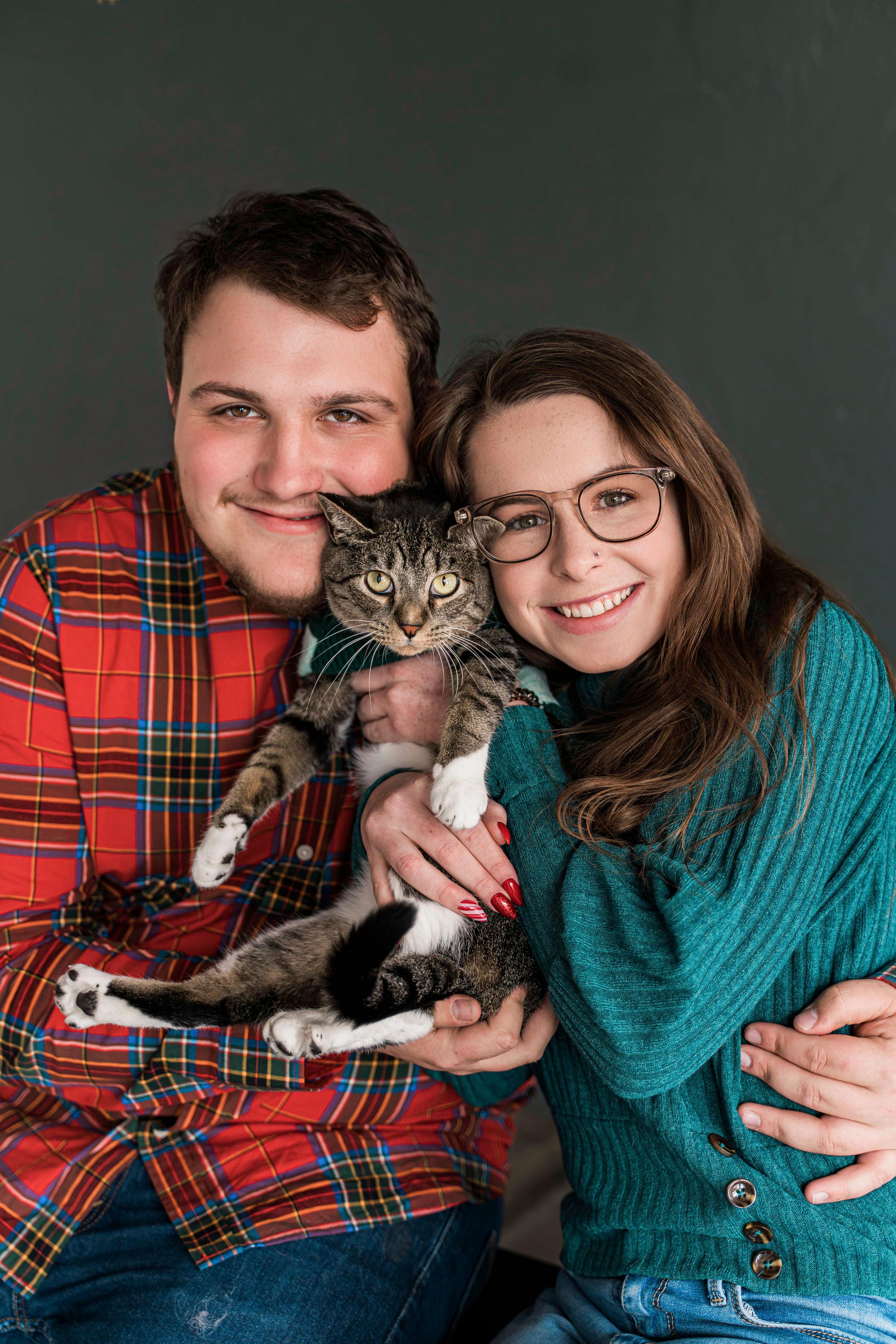Couple in festive sweaters poses with their gray tabby cat against dark green background.