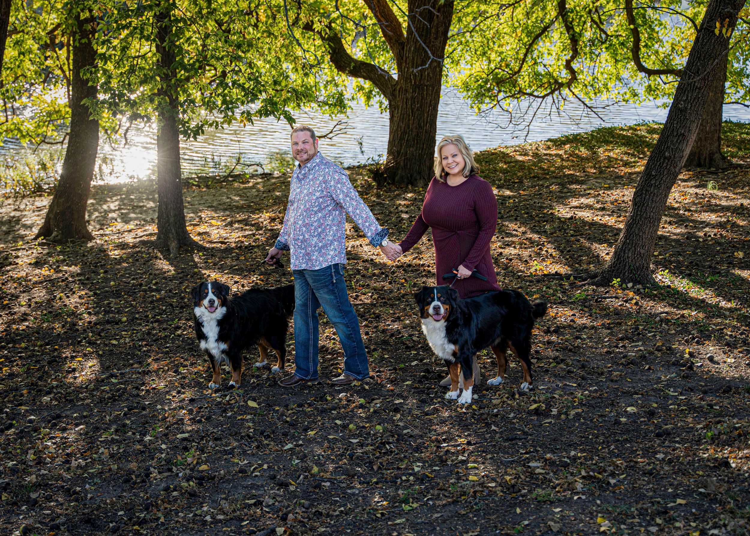 Couple walk their black and white dogs on a leaf-covered path under sunlit trees in a park.