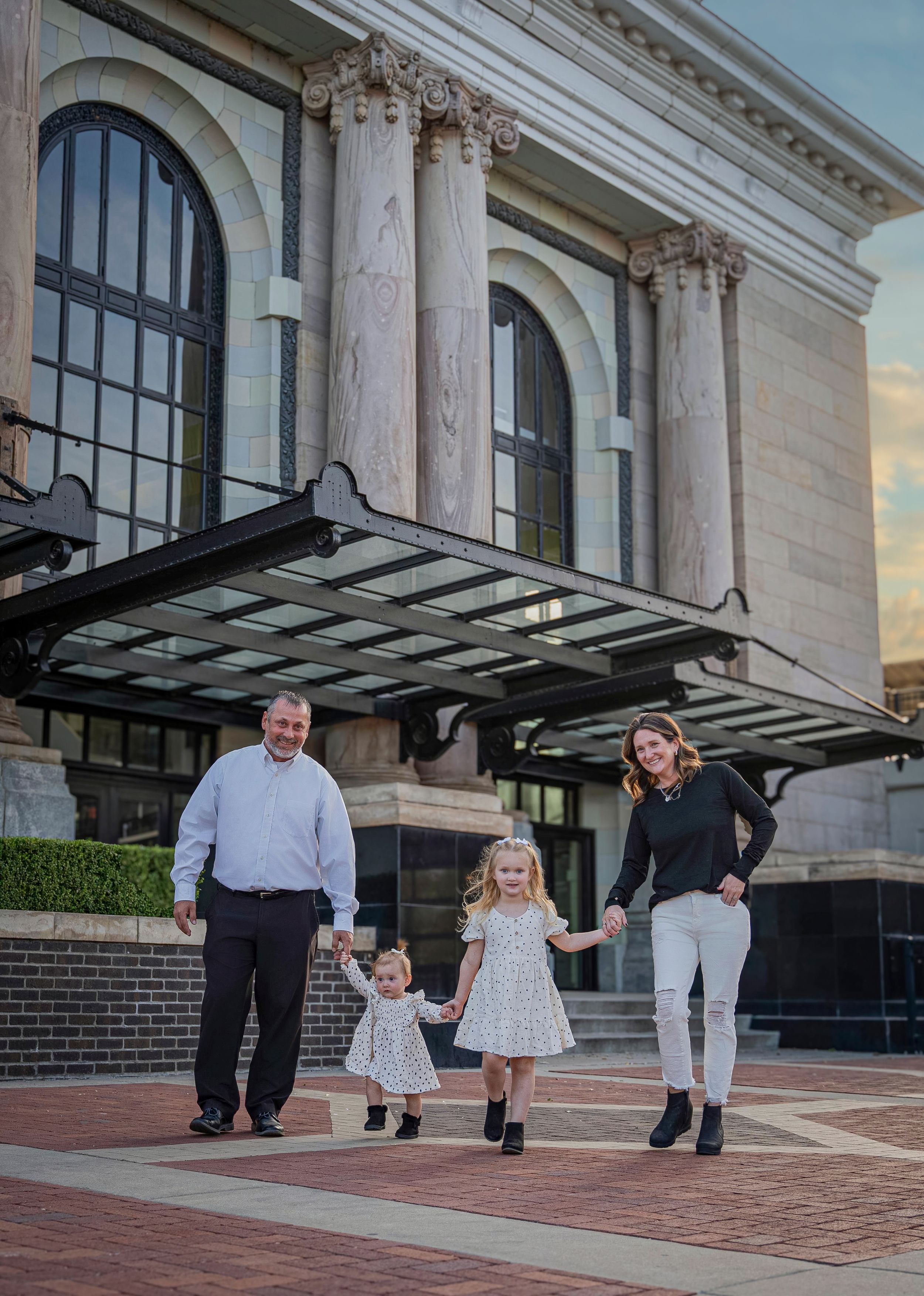 A family holds hands while walking together in front of a historic building with classical columns and glass awning.