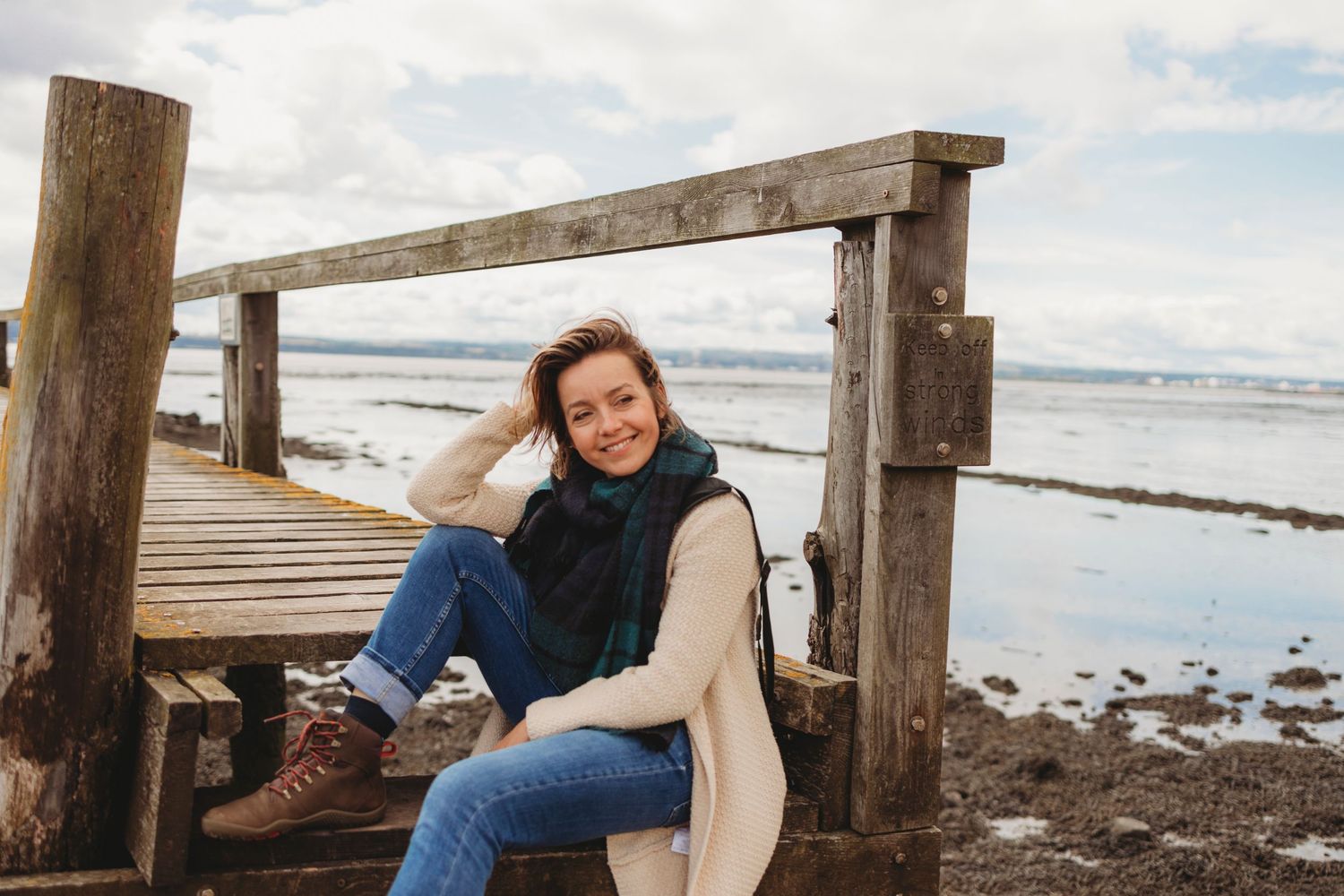 Someone in a cream sweater and jeans sits on a wooden dock by the beach on a cloudy day.