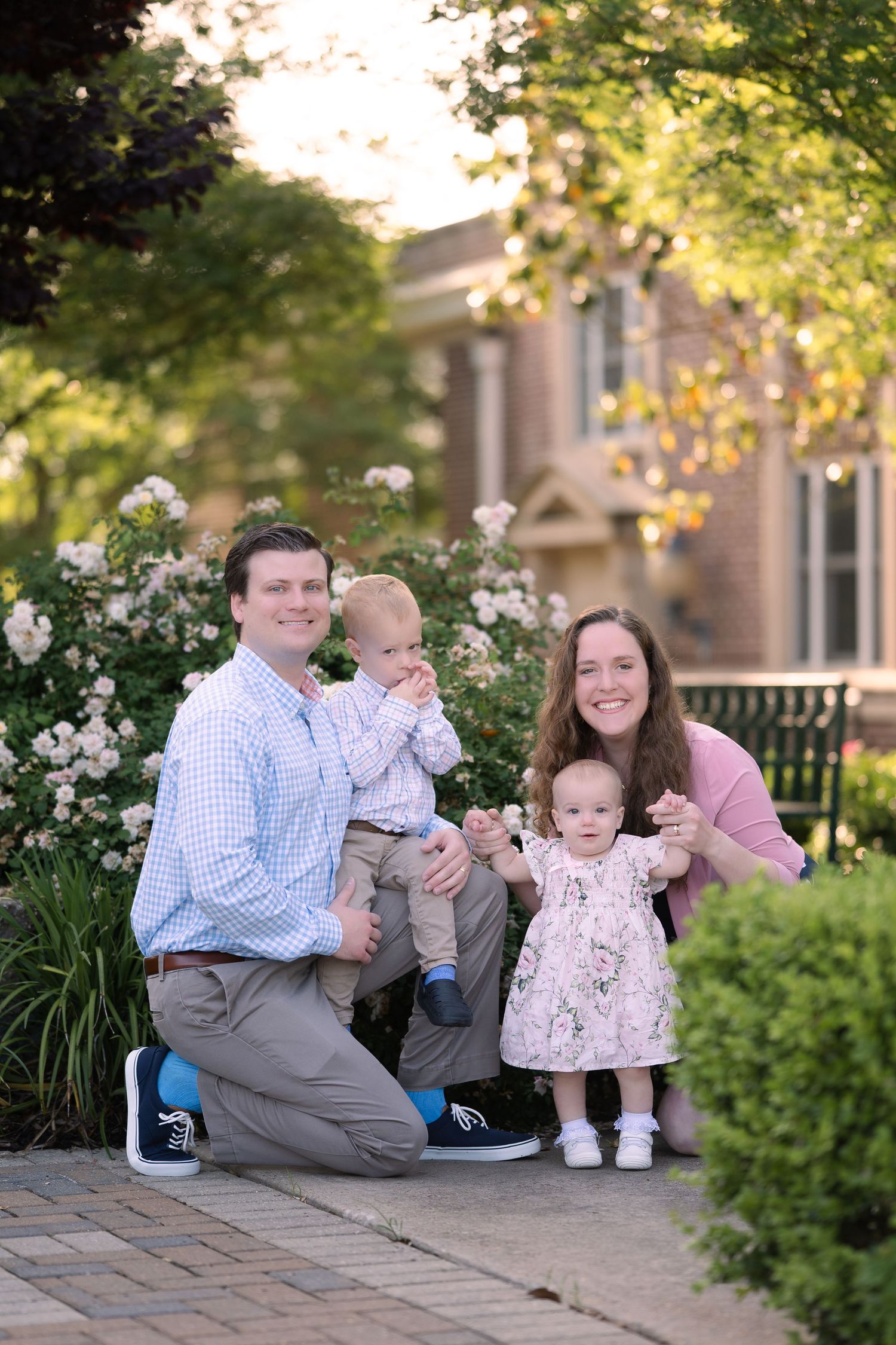 Family poses together on a brick pathway in front of flowering bushes and a building during golden hour.