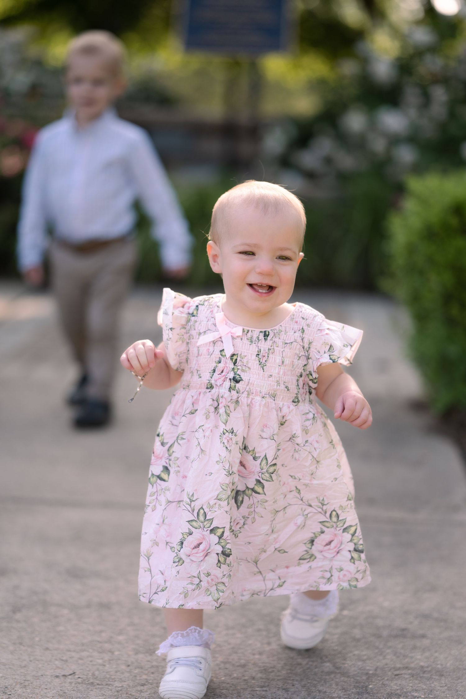 A small child in a floral pink dress and white socks runs joyfully down a path on a sunny day.