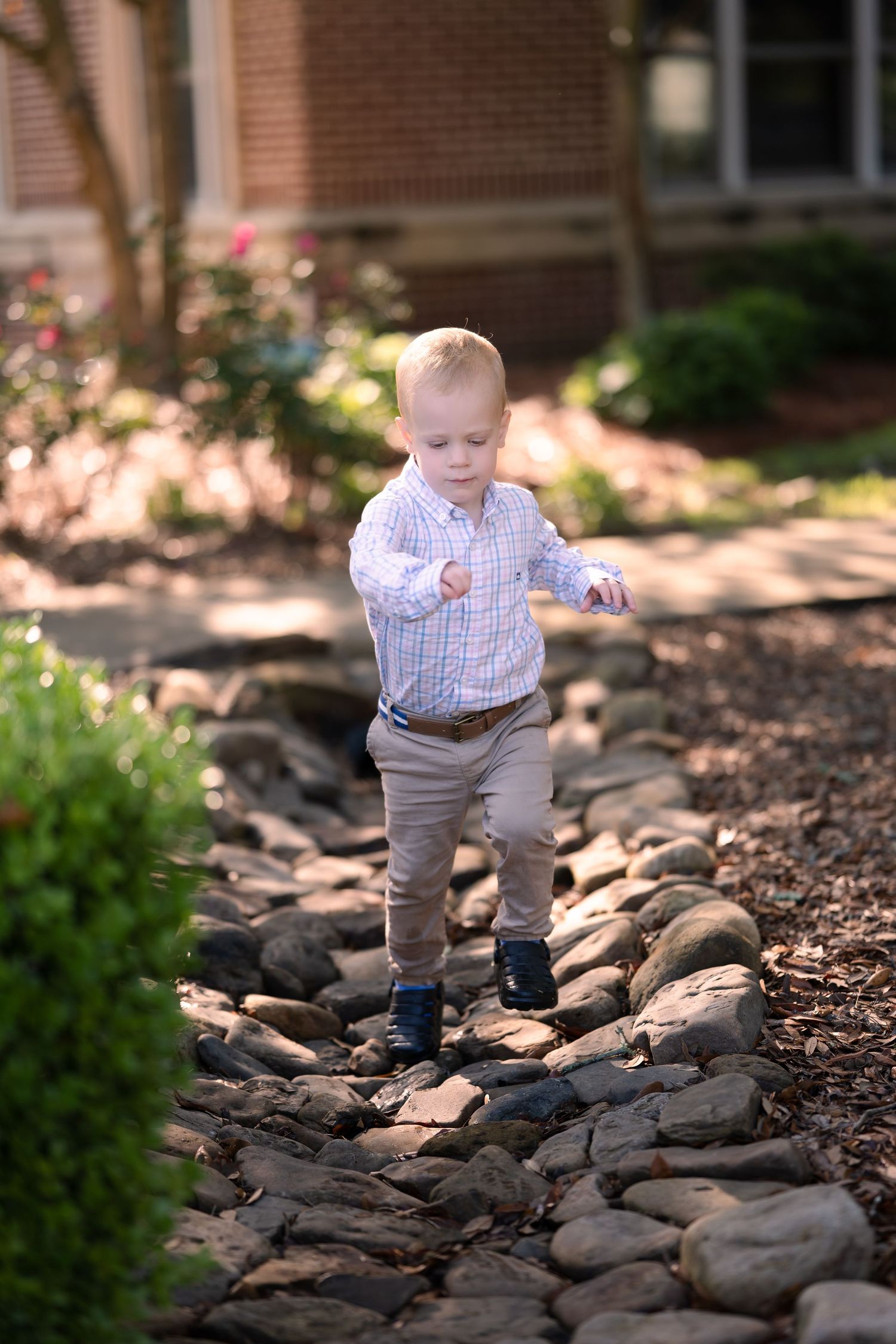 Small child carefully walks along rocky garden path wearing checkered shirt and khaki pants on sunny day.