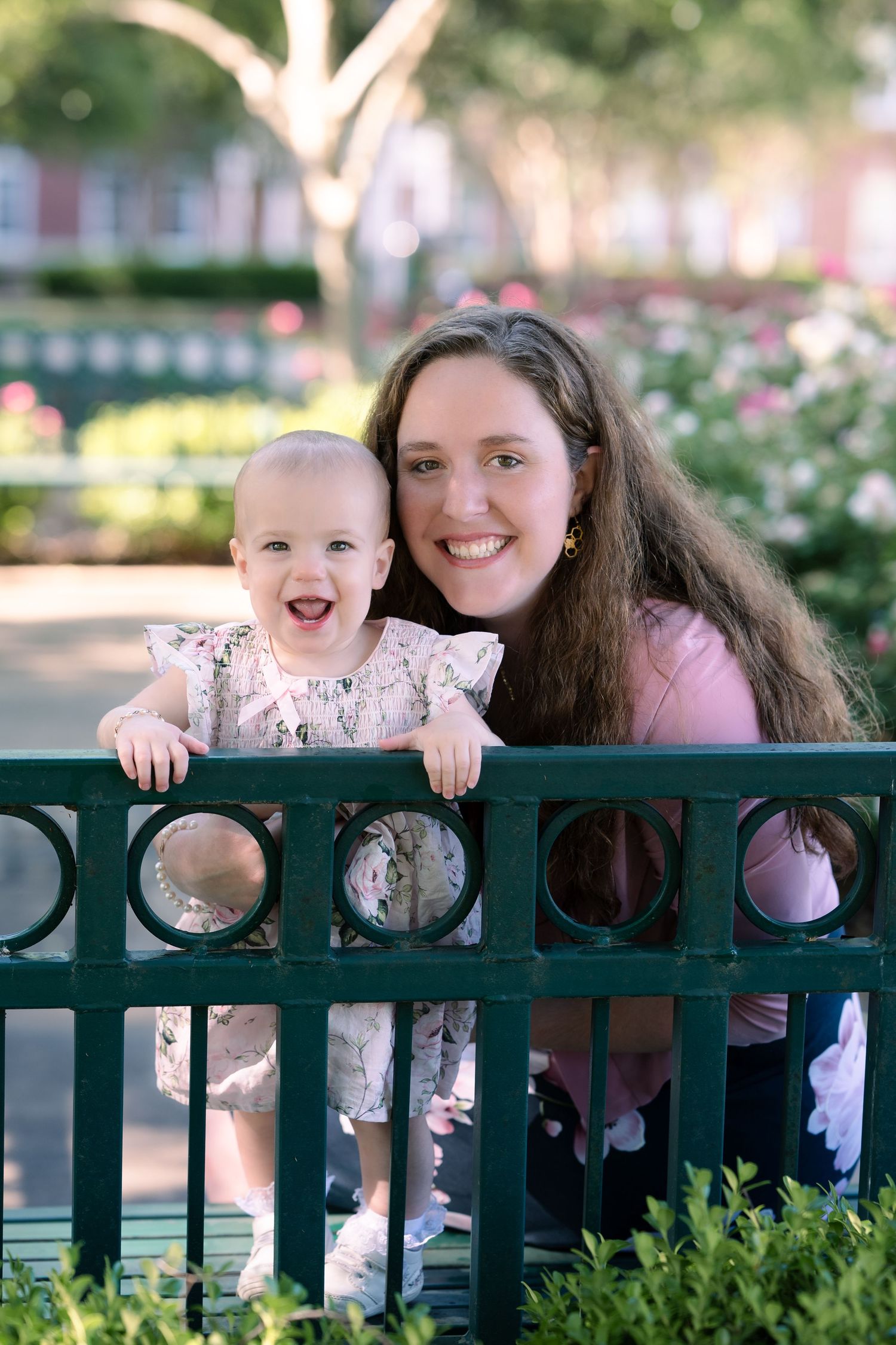 A happy adult and young baby share a joyful moment while standing by a decorative green railing in a garden setting.
