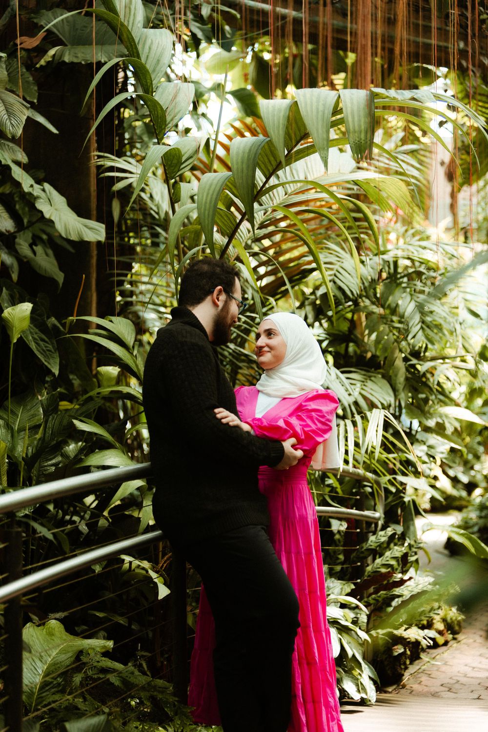 Couple posing among lush tropical plants with vibrant pink dress creating dramatic contrast.