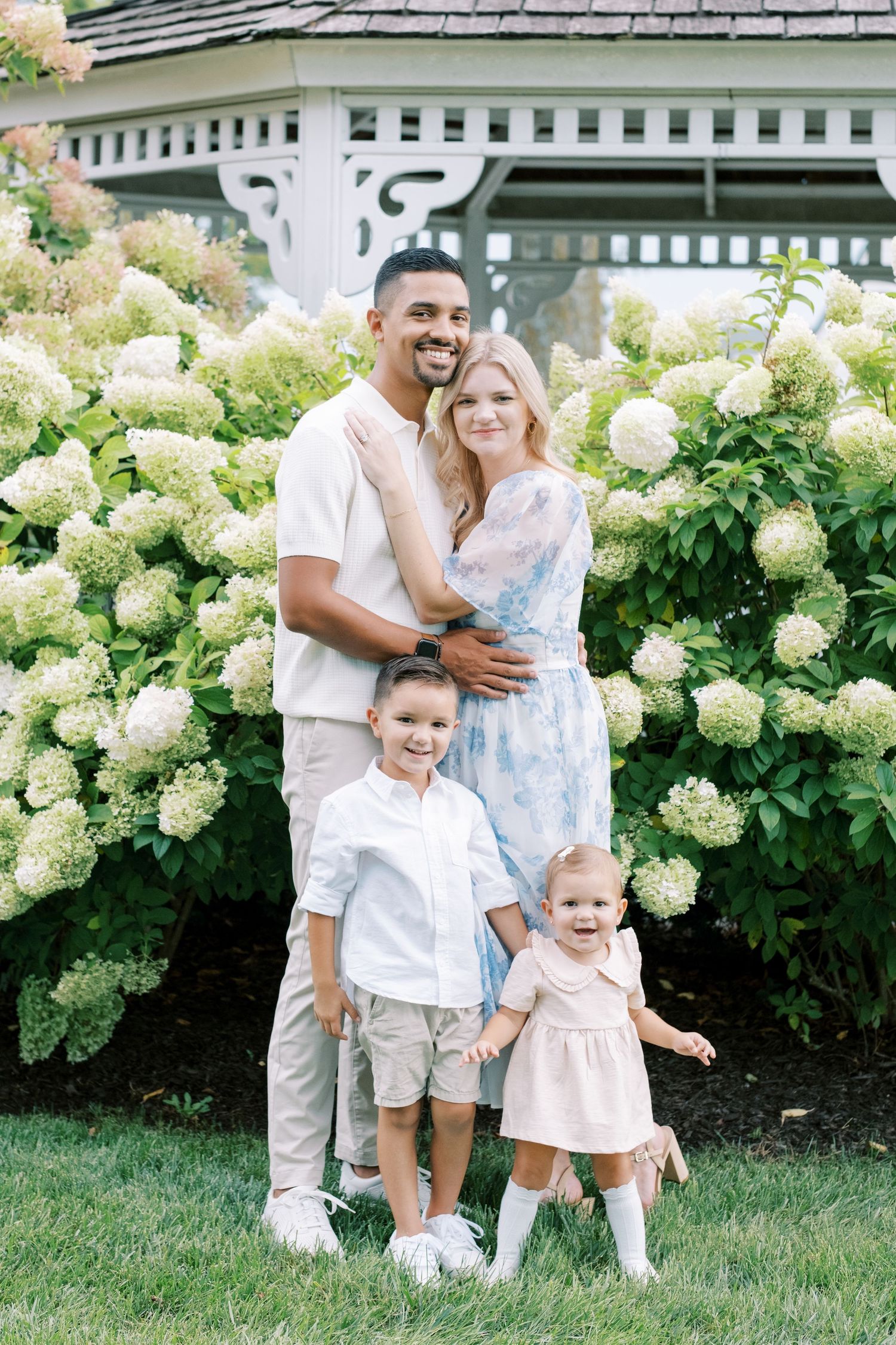 A beautiful family portrait in light summer clothing against blooming hydrangea bushes and white gazebo.