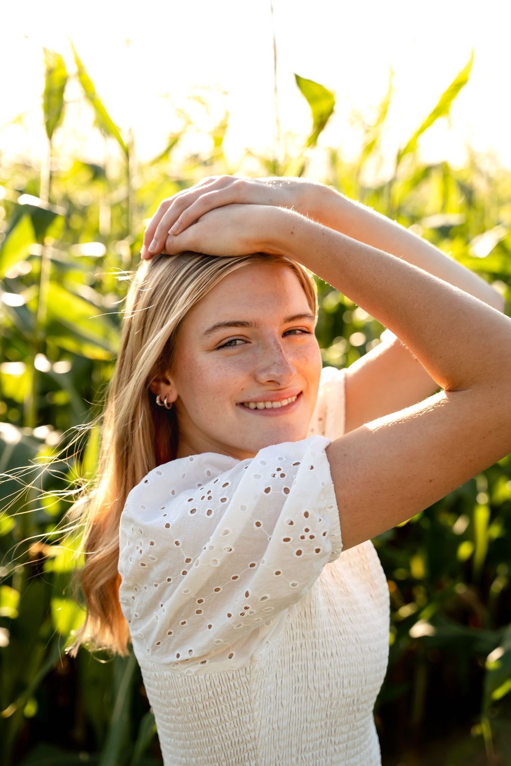 A person in a white eyelet dress poses in golden sunlight near tall corn stalks.