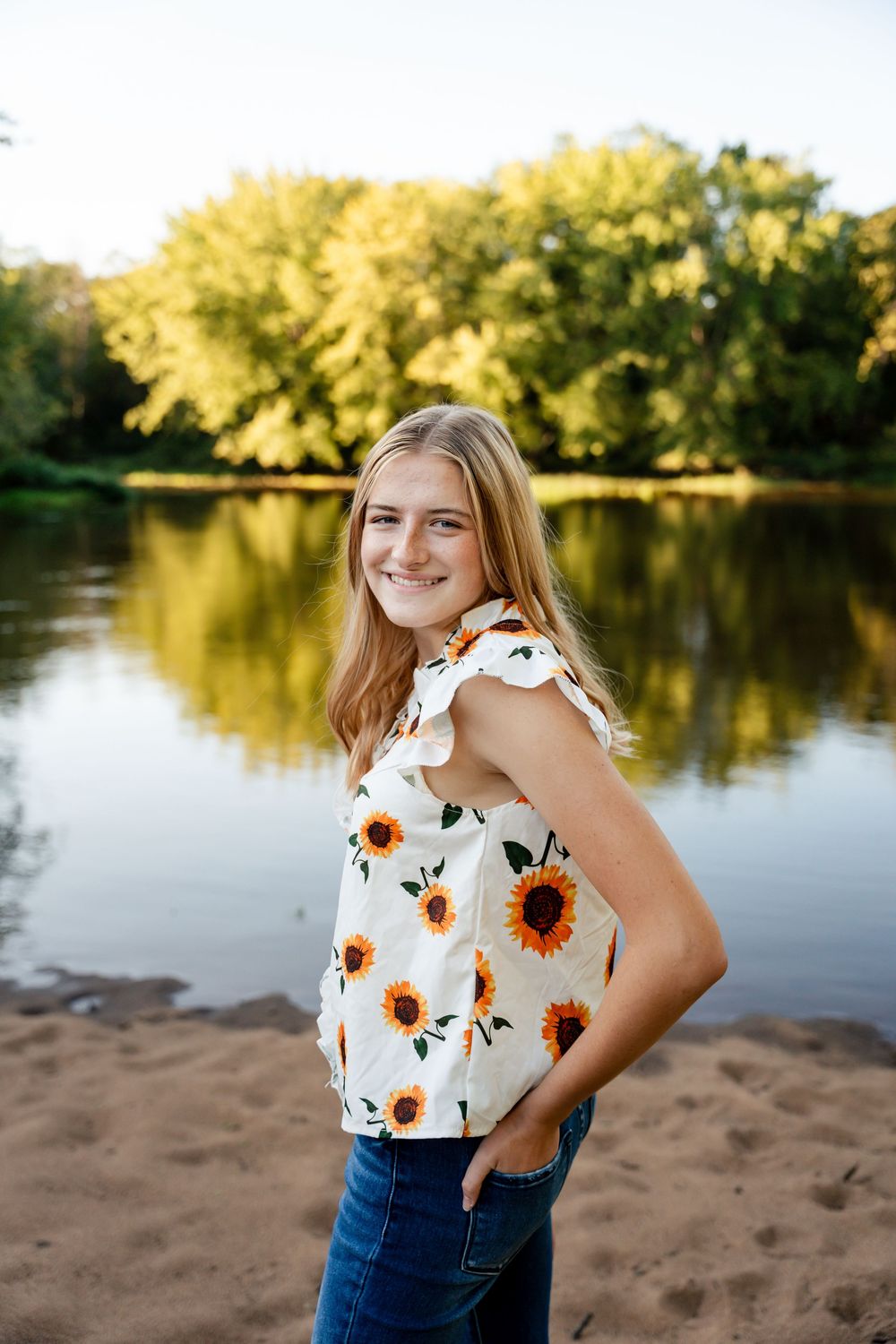 A person in a sunflower print top poses by a serene lake at sunset.
