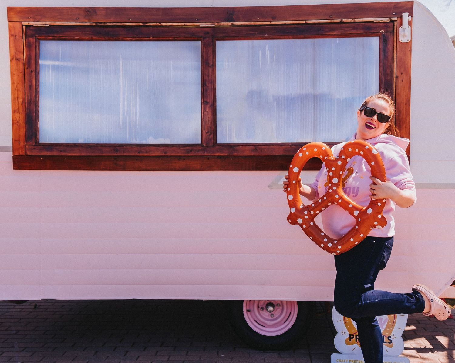 Person in sunglasses poses with a heart-shaped pretzel next to a white camper van with wooden window frame. Pretzel food truck in Maine.